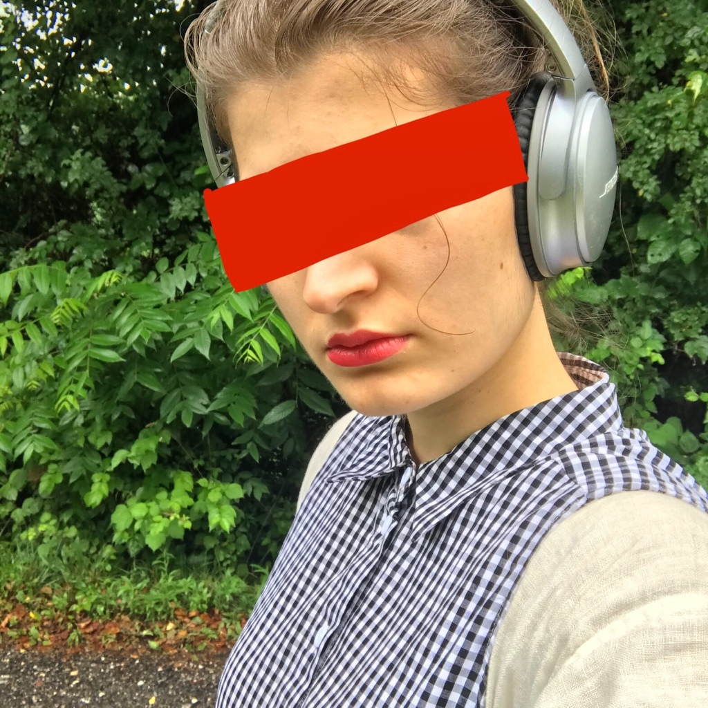 The author is a fair-skinned person wearing headphones.  Their lips are painted red, and their eyes are covered by a big red block to obscure their identity.  They are wearing a white long sleeved shirt underneath a black and white gingham collared, sleeveless button down that is fully buttoned.  They stand against a backdrop of green leaves.