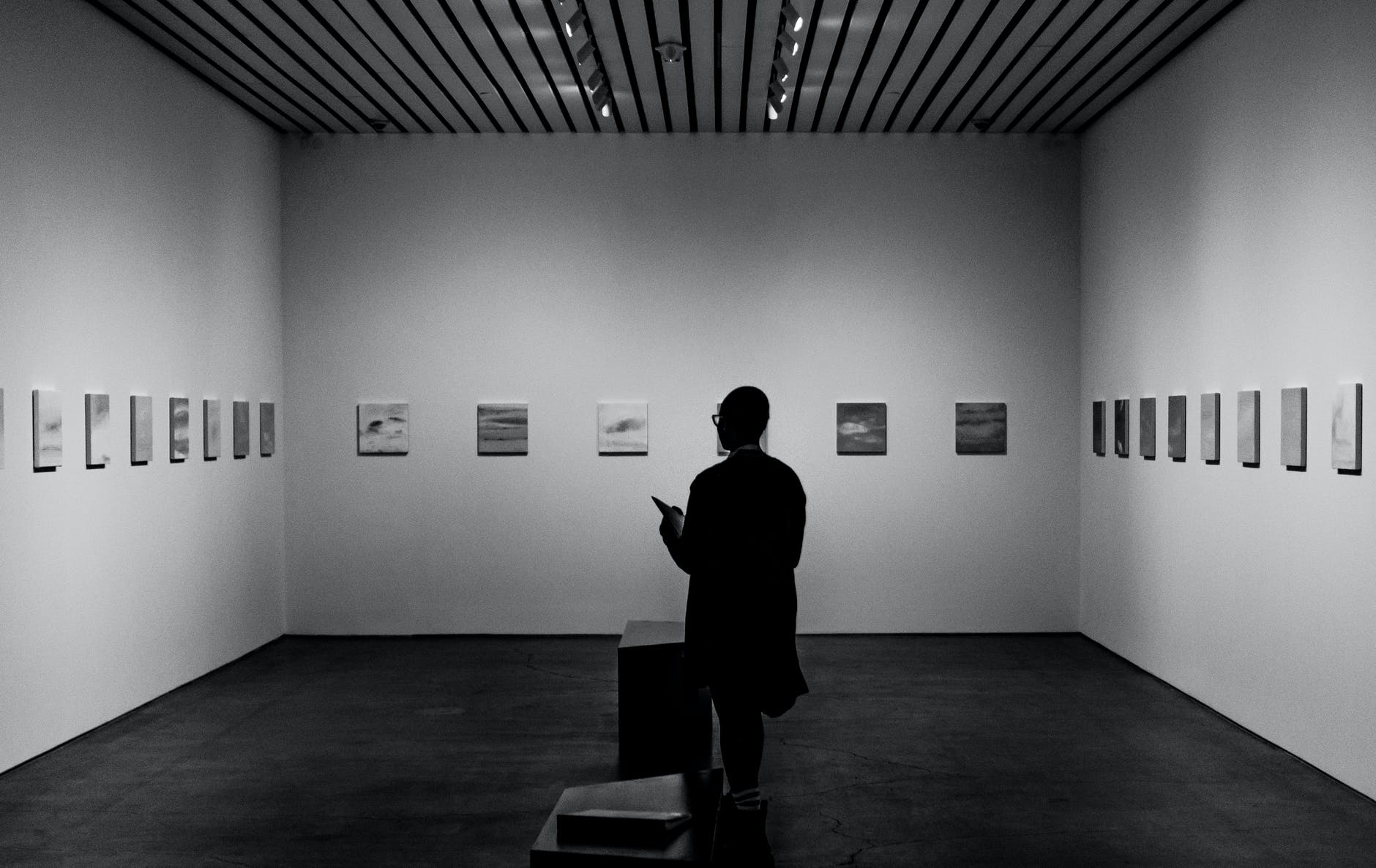 A shadowed person wearing glasses stands in an empty room.  The walls are white, and many pictures hang in the wall.  A gallery.