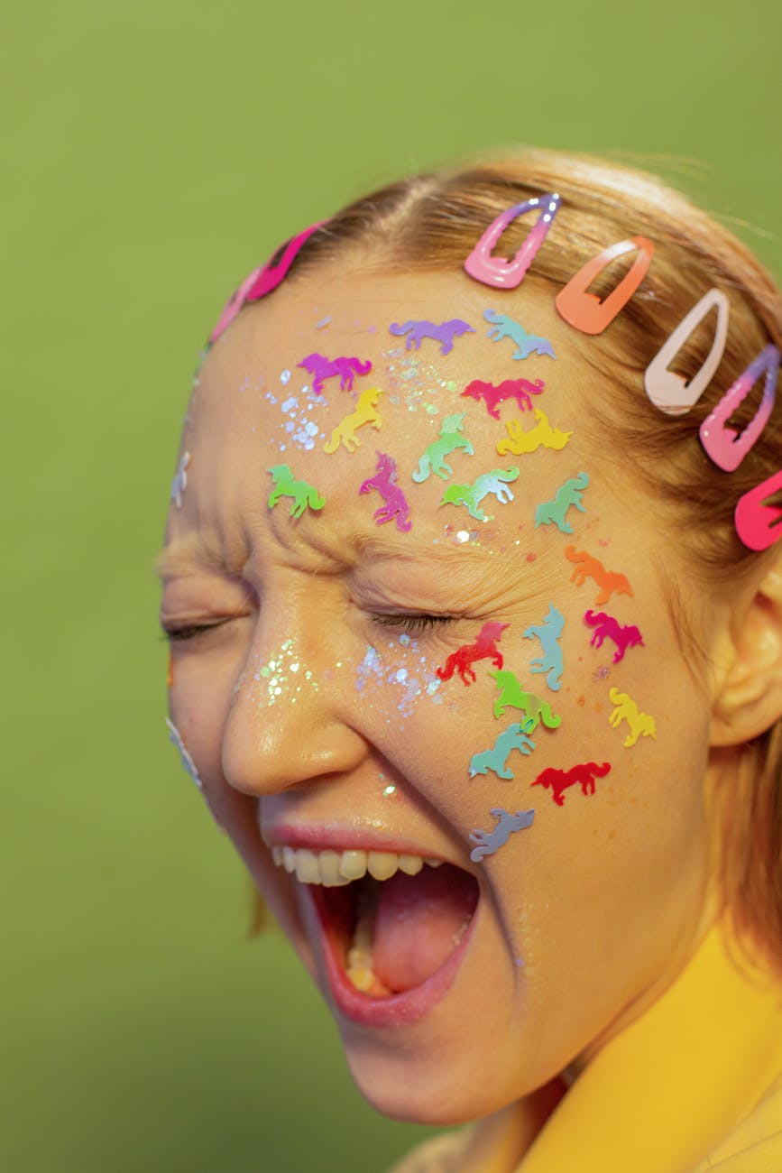 A light skinned femme with blonde, chin length hair stands against a green background.  There are pink barrettes in various shades pinning their hair back.  Stickers of animals in various colors bedazzle their face.  Their eyes are closed, brow furrowed, and their mouth is open as if yelling.  They are wearing a bright yellow shirt.